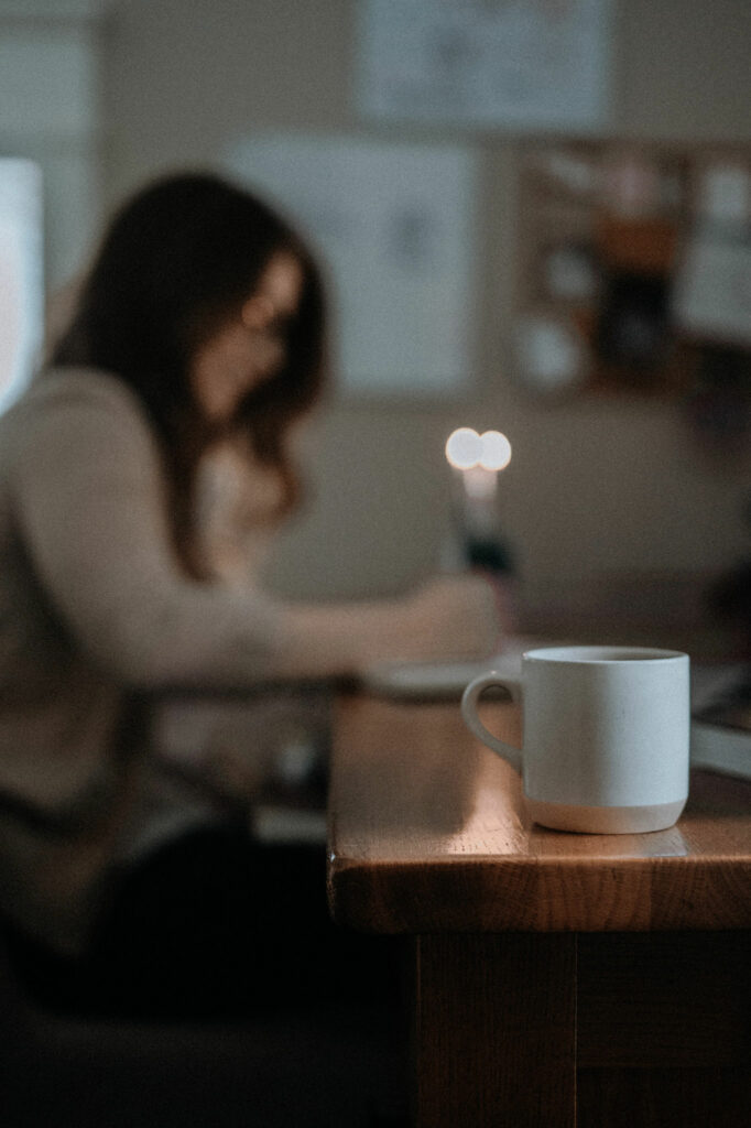 coffee cup on edge of desk. girl at the desk writing with candle burning in background
