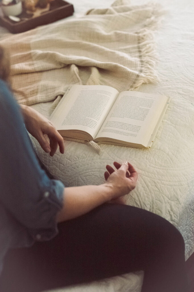 side of girl wearing blue sitting on edge of bed with an open book on the bed in front of her