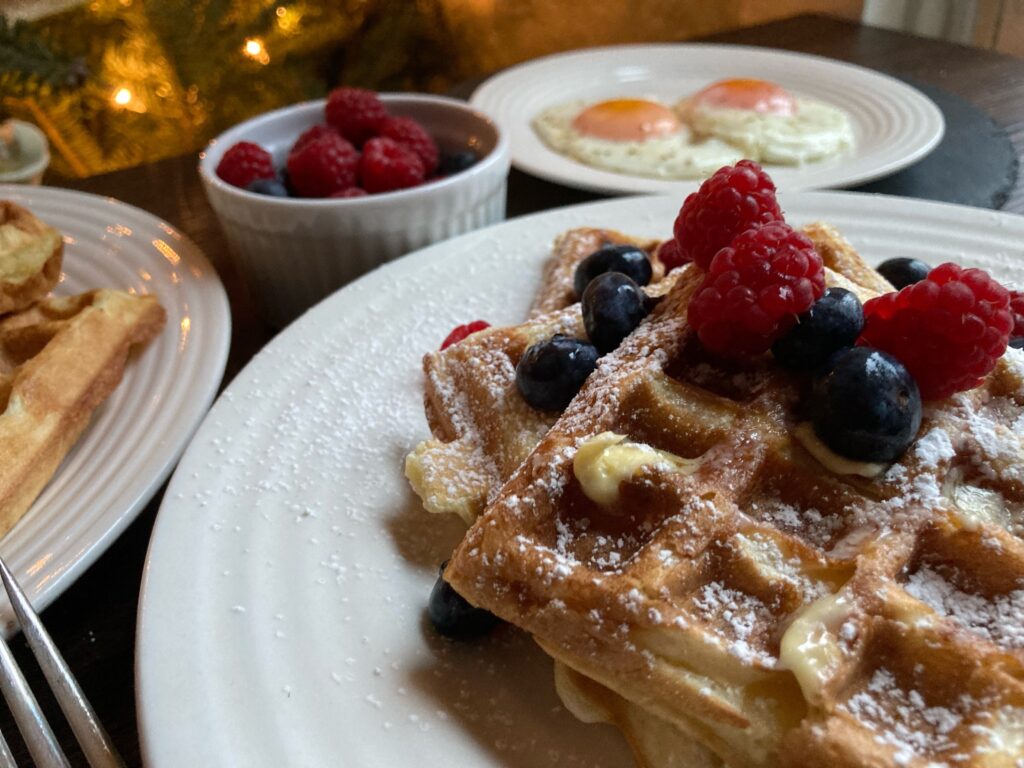 side shot of sourdough discard waffles covered in berries and powdered sugar with eggs in the background