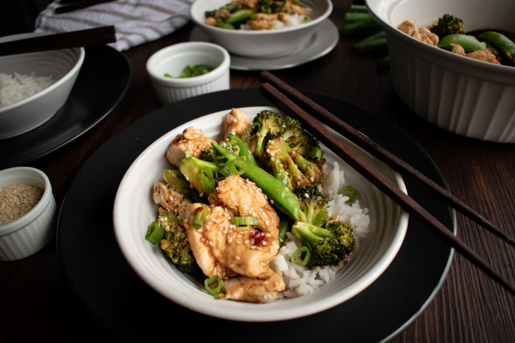 Sesame chicken in a bowl with chopsticks and other ingredients surrounding it