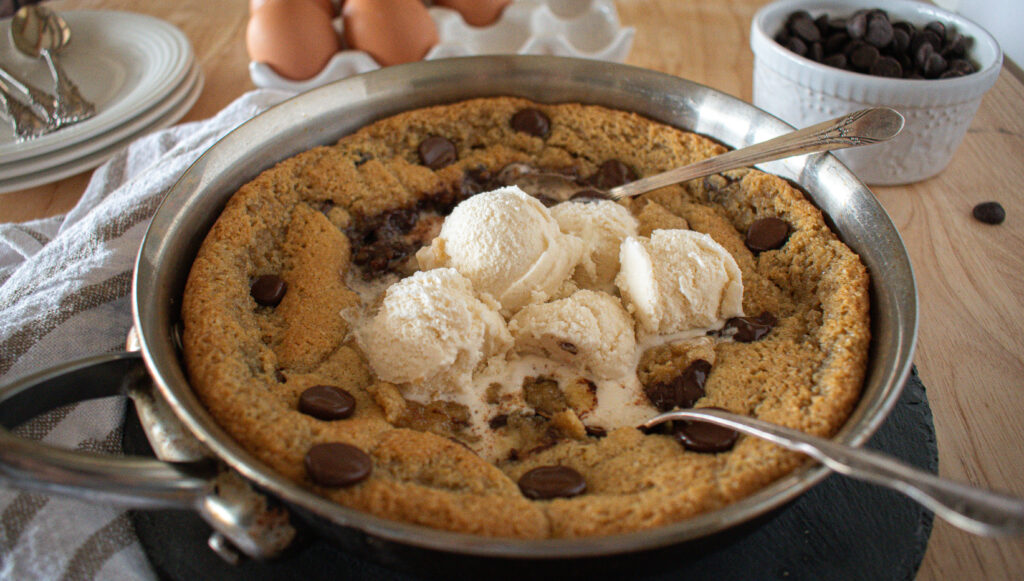 a chocolate chip cookie made in a skillet with spoons scooped inside and vanilla ice cream melting on top. 